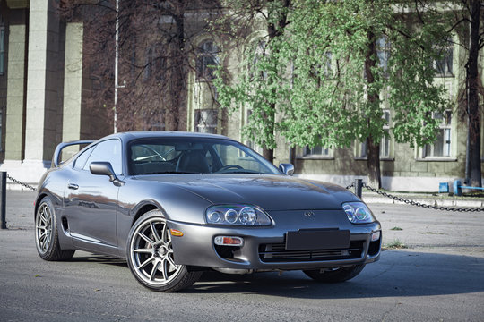 A very rare Japanese sports car in the back of a Toyota Supra coupe in gray with a high spoiler on the road in the city near trees with yellow leaves.