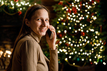 A young pretty girl is talking on a mobile phone, writing a message, using the Internet on the background of a Christmas tree and a fireplace.