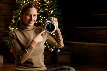 A young girl holding a watch 5 minutes before the new year on the background of many lights, a Christmas tree and a fireplace. Peal of bells.