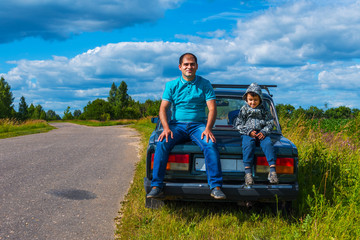 dad and son sitting on the back of a car near the road. dad and son travel by car