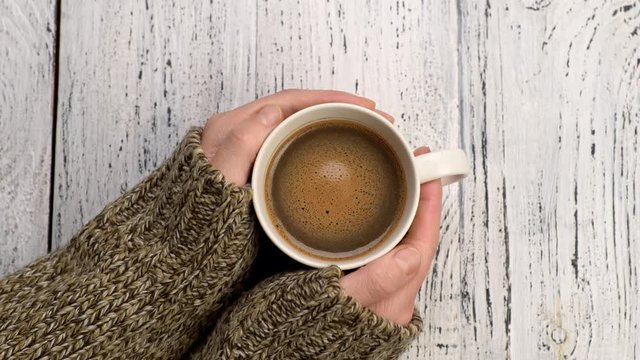 Female hands holding cup of hot coffee on old white wooden background. Coffee foam swirls in white cup. Winter concept