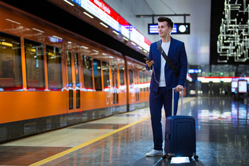 Young stylish handsome man in suit with suitcase standing on metro station holding smart phone in...