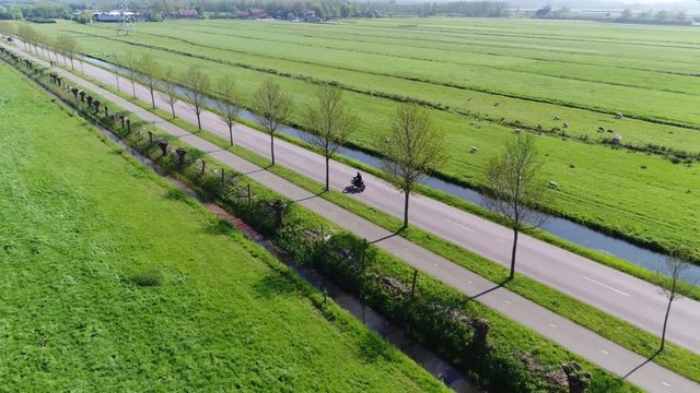Aerial following motor cyclist driving on road in rural area beautiful grass landscape with meadow and pasture left and right of straight countryside way also showing a bikeway on side of treeline 4k