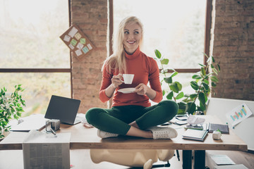 Full body photo of happy positive woman company owner sit on table crossed legs rest relax hold coffee cup drink cappuccino wear red turtleneck green pants trousers in messy office loft - obrazy, fototapety, plakaty