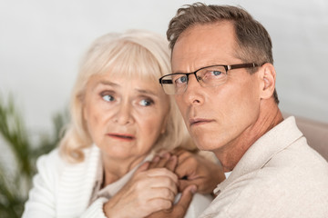 selective focus of sick senior man in glasses holding hands with wife