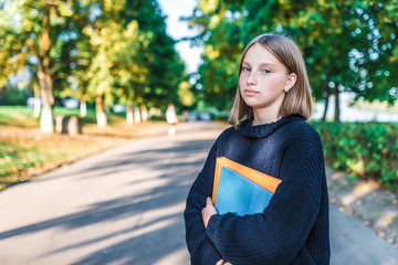 Girl teenager schoolgirl, summer city stands in sweater, hands of textbooks, notebooks and notes, smiling, resting on vacation, background trees. Free space text. Preparing school institute college.