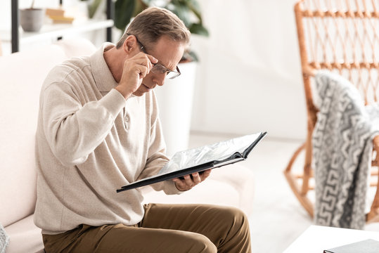 senior man touching glasses and holding photo album while sitting on sofa