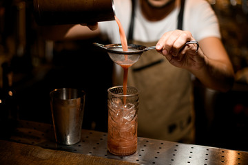 Fototapeta na wymiar Professional male bartender pouring a red alcoholic drink from steel shaker through the sieve