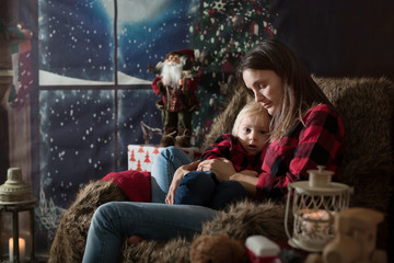 Fototapeta na wymiar Beautiful young mother, hugging her toddler boy, sitting in cozy chair on Christmas