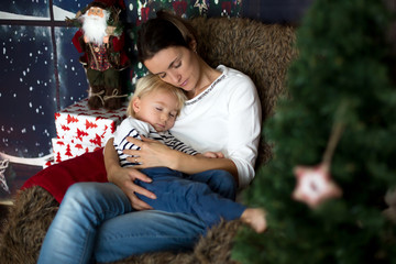 Beautiful young mother, hugging her toddler boy, sitting in cozy chair on Christmas