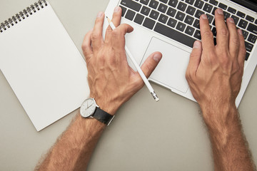 cropped view of man typing on laptop while holding pencil near notebook isolated on grey