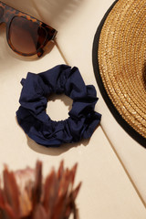 Subject shot of a shimmering blue textile hair band with a textured piping. The scrunchie is isolated on the beige fabric surface and surrounded with a brown straw hat, sunglasses and tropic flowers. 