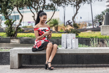 Adult ethnic woman using smartphone while sitting on bench beside paper bags in downtown