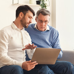 Son Teaching Senior Father Computer Literacy Sitting On Sofa Indoor