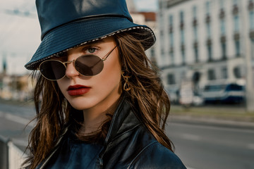 Outdoor close up fashion portrait of young elegant model, woman wearing trendy faux leather bucket hat, round sunglasses, earrings, posing in street of European city. Copy, empty space for text