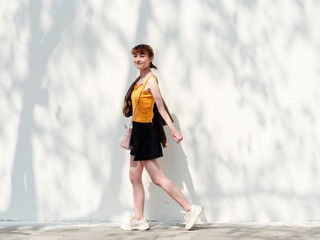 Portrait of happy Chinese girl in yellow halter top and short skirt posing on sunny street with white wall background and tree shadows, carefree young lady enjoy her leisure time.