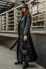 Outdoor full-length fashion portrait of young confident woman wearing long black leather trench...