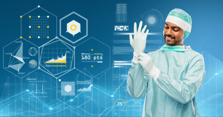 Fototapeta na wymiar medicine, surgery and people concept - smiling indian male doctor or surgeon in protective wear putting glove over virtual charts projection on blue background