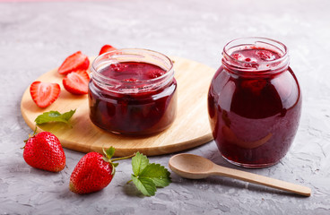 Strawberry jam in a glass jar with berries and leaves on gray concrete background, side view.