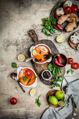 Tom Yum soup with shrimps in bowls on shabby beige background with limes, chili pepper, ginger root and mushrooms, ingredients for cooking, copy space
