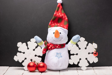 Cheerful snowman toy in a red hat and scarf with foam snowflakes on the background of black chalk...