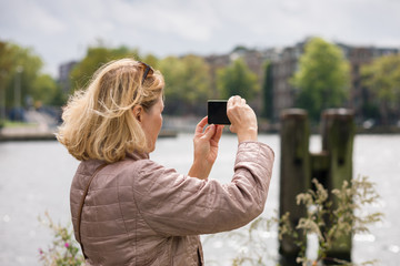 An elderly woman photographs the sights of the city on a mobile phone, walking through the streets of the summer city. Concept of active lifestyle of elderly people.