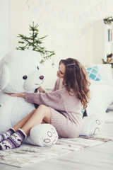 Beautiful caucasian woman in sweater sitting on floor with teddy bear in cozy room