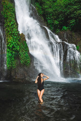 Young slim brunette woman with curly hair enjoying in lagoon of huge tropical waterfall Banyumala in Bali. Wearing in black swimsuit. Happy vacations in Indonesia. Wanderlust travel concept.