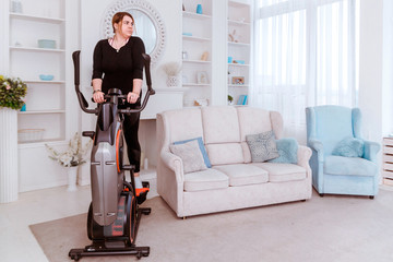 Model - a fat woman, trying to lose weight and at home is engaged in an elliptical trainer