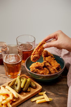 Cropped View Of Woman Eating Delicious Chicken Nuggets, French Fries And Gherkins Near Glasses Of Beer On Wooden Table Isolated On Grey