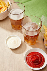 glasses of beer, french fries, ketchup and mayonnaise on wooden table on grey background
