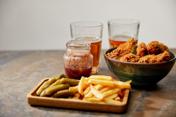 delicious chicken nuggets, french fries and gherkins near glasses of beer on stone surface isolated on grey