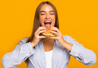 Hungry Woman Biting Burger Standing Over Yellow Background