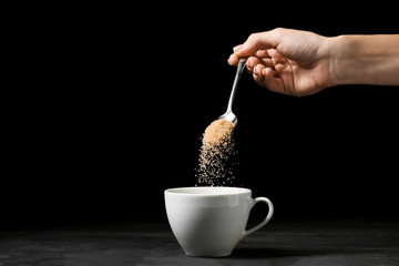 Woman pouring sugar from spoon into cup on dark background