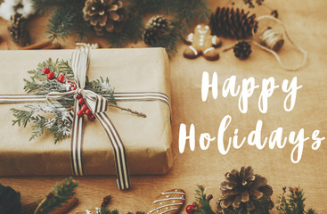 Happy Holidays text sign on stylish rustic christmas gift, pine branches, cones, gingerbread cookies, cinnamon, anise on rural wooden table, Season's greeting card