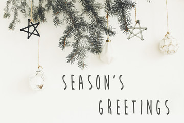 Season's greetings text sign on stylish christmas branches with glass modern ornaments hanging on white wall. Creative christmas festive decor. Seasons greeting card © sonyachny