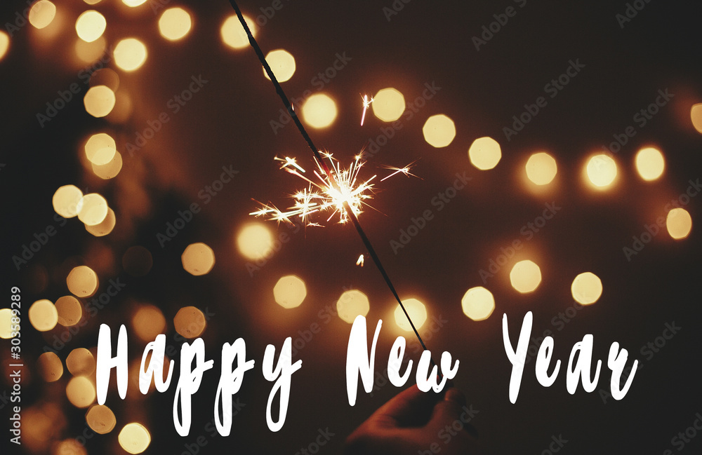 Wall mural Happy New Year text sign on glowing sparkler in hand on background of golden christmas tree lights in dark festive room. Fireworks burning. Seasons greeting card