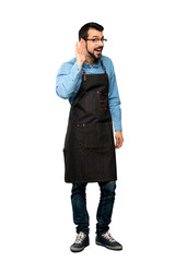 Fototapeta premium Full-length shot of Man with apron listening to something by putting hand on the ear over isolated white background