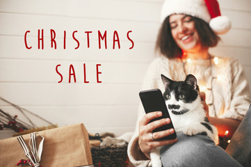 Christmas Sale text sign on cat looking at phone screen with funny emotions and sitting on happy girl legs in christmas lights. Special discount offer. Holiday shopping online