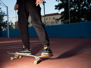 male skateboarder skating in action in a skatepark with long black pants and sneakers with sports field background and buildings