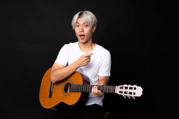 Young asian man with guitar over black background surprised and pointing finger to the side