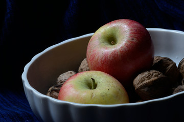 apple and whole walnuts in a white fruit bowl against a blue background