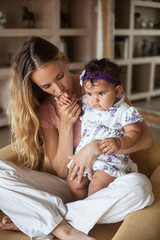 Obraz na płótnie Canvas beautiful young blonde mom holds in her arms her toddler daughter. A woman is sitting on a sofa indoors and kissing hand of girl. Multiracial family.