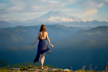 Naklejka premium Piękna dziewczyna w lekkiej letniej sukience w górach Kaukazu, podziwiająca zachód słońca i ciesząca się wolnością i czystym górskim powietrzem.