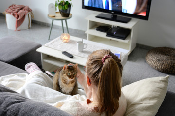 Domestic life with pet. A young woman is sitting on the couch with her cat on her lap in the living room. She watches TV while stroking her cat. Woman binge watching tv via online streaming platform.