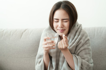 Asian Cute of  girl having headache and high temperature from illness holding pill of medicine and water sitting on sofa at home, Health and illness concepts