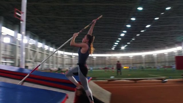 Pole vaulting indoors - a young woman with ponytail jumping over the bar and falling down