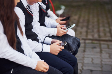 Close up view of school kids in uniform that sits on the bench with smartphones
