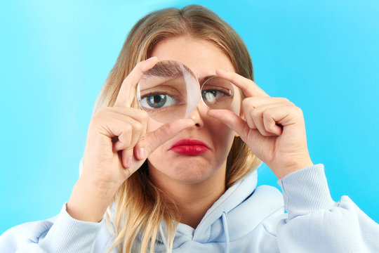 Fototapeta Young upset woman looking at the camera through magnifier big eye closeup isolated on blue. Bad vision, search concept