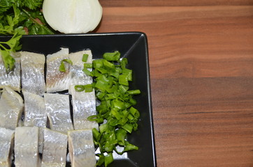 On the table is a black rectangular plate with Norwegian herring fillet and chopped onion and parsley sprigs.